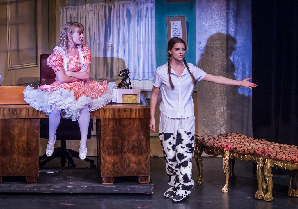Gypsy — a girl in an orange gingham dress with white petticoat sits cross-armed on a wooden desk before a teal-walled office set with curtained windows, while Louise in a white shirt and black-and-white cow-print pajama pants gestures with one arm extended toward her