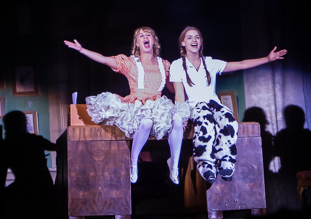 Gypsy — the gingham-dress girl and Louise in her cow-print pajamas sit side by side on a wooden desk with arms spread wide and mouths open in full-voiced song, the audience silhouetted in the foreground against the teal office set