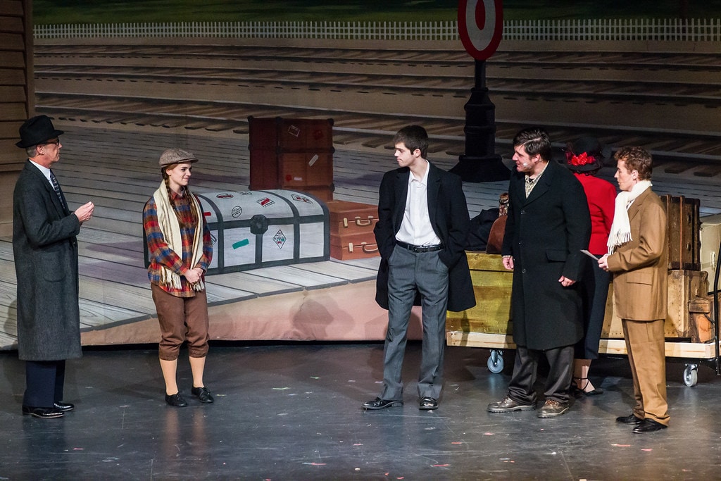 Gypsy — five performers in 1930s winter coats and hats stand before a painted train station backdrop with vintage luggage trunks: a man in a black hat at left addresses a young woman in a plaid jacket and scarf, while two men in dark overcoats and a young man in a tan suit look on at right