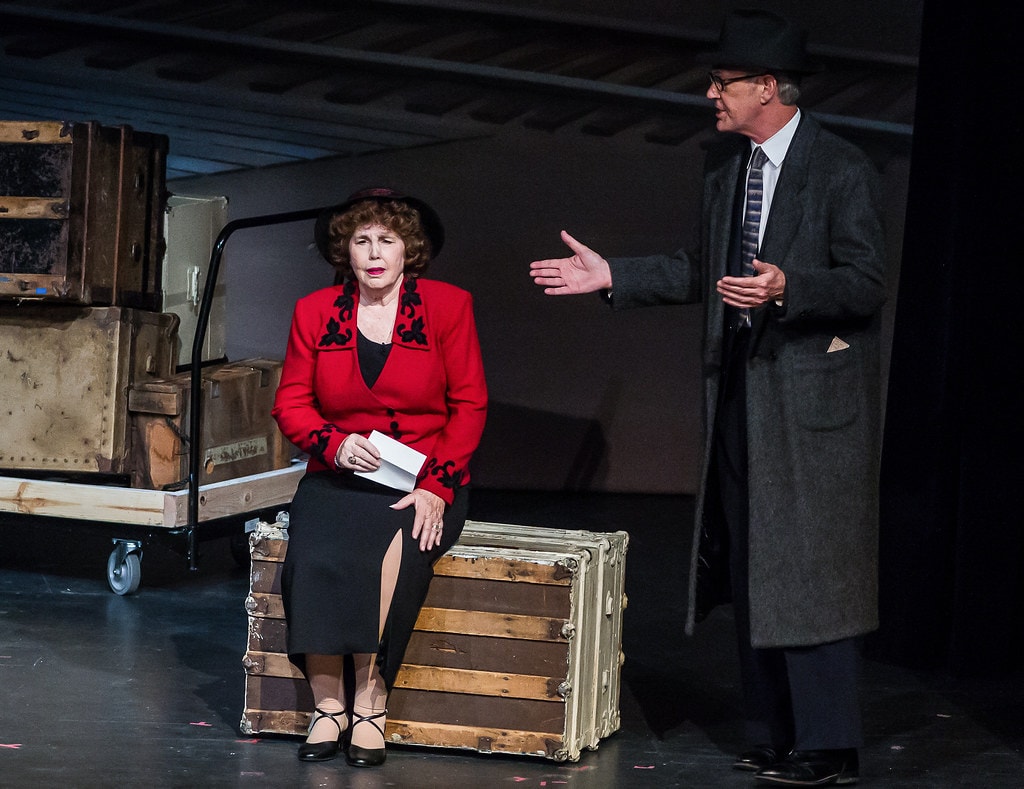 Gypsy — Rose in a red blazer with black floral trim sits on a vintage steamer trunk holding an envelope as a man in a grey overcoat and black hat extends a hand toward her before a dark backstage backdrop with a luggage cart stacked with wooden crates
