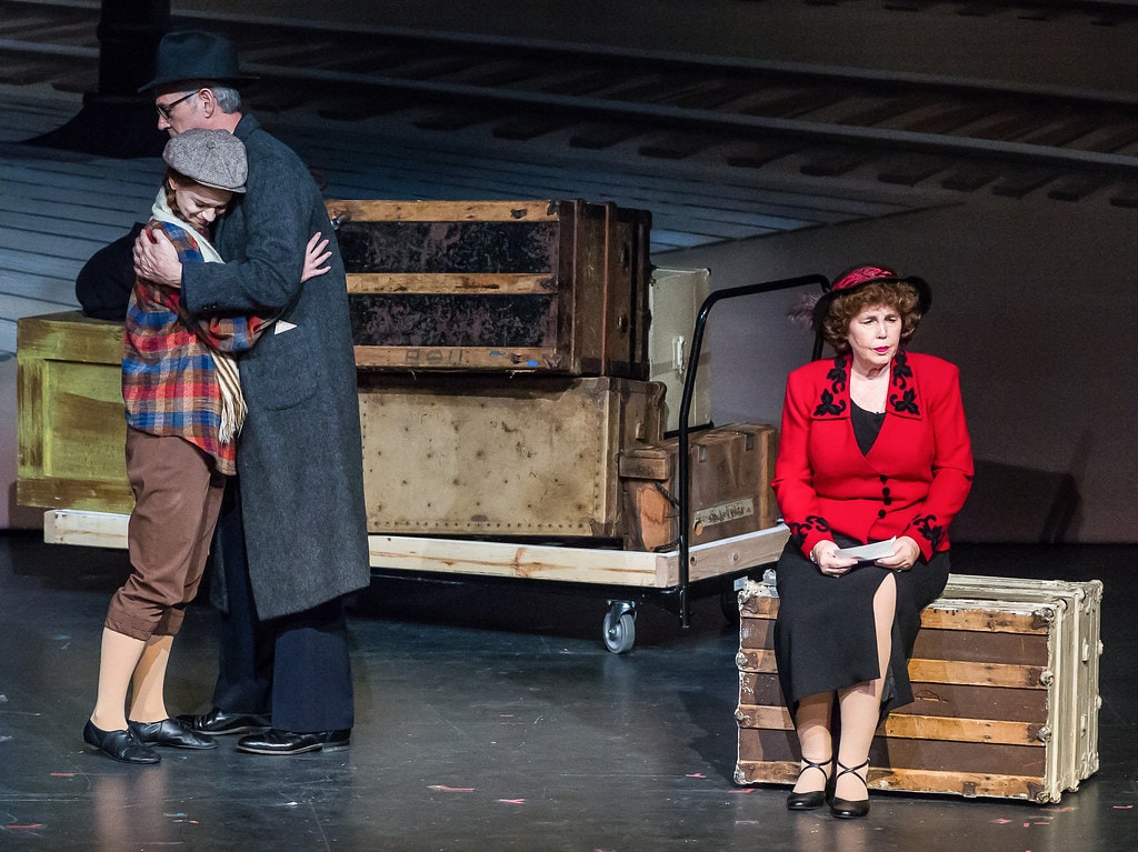 Gypsy — the young woman in the plaid jacket and scarf embraces the man in the grey overcoat in a farewell hug at left as Rose in her red blazer sits on the steamer trunk holding her envelope at right, the luggage cart and crates visible behind them