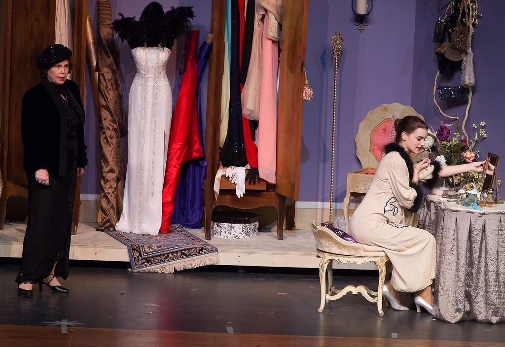 Gypsy — Mama Rose's boudoir confrontation: an older woman in a black sequined dress and beret stands at left surveying the room as Louise in a cream fur-trimmed robe sits at a gilded vanity table applying makeup, the elaborate costume rack with a white beaded gown and colorful silks visible between them on the lavender-walled set