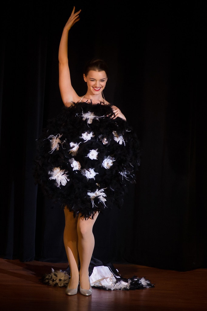 Gypsy — a performer in a dramatic black feathered ball gown with white floral accents raises one arm high and smiles against a black backdrop