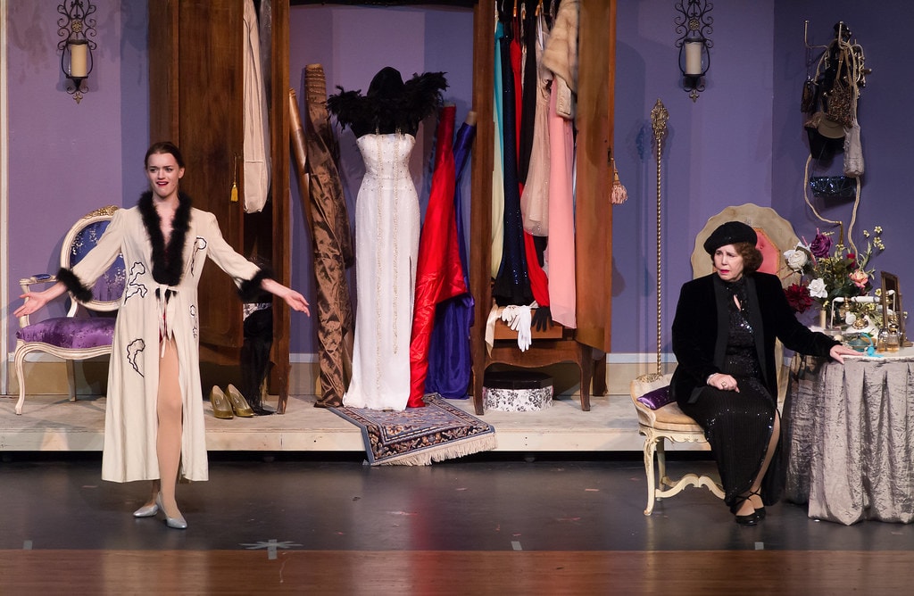 Gypsy — Louise in a cream fur-trimmed robe gestures with arms wide as Rose in a black sequined dress sits at the vanity table before the purple-walled dressing room set with a rack of gowns and a Persian rug