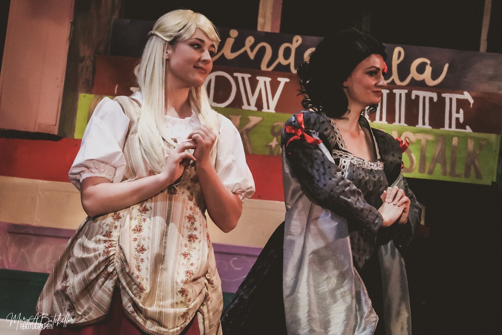 Into the Woods — Rapunzel in a blonde wig and Snow White in a dark gown stand before the colorful storybook backdrop