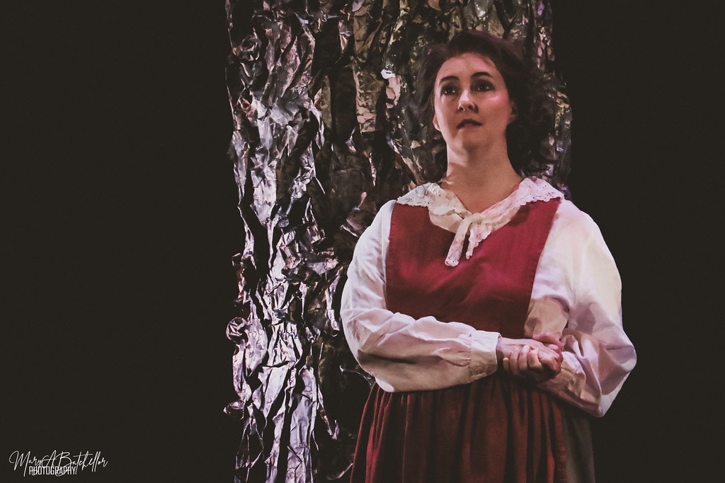 Into the Woods — Little Red Riding Hood in a red dress and white blouse sings a solo before a silver foil tree backdrop