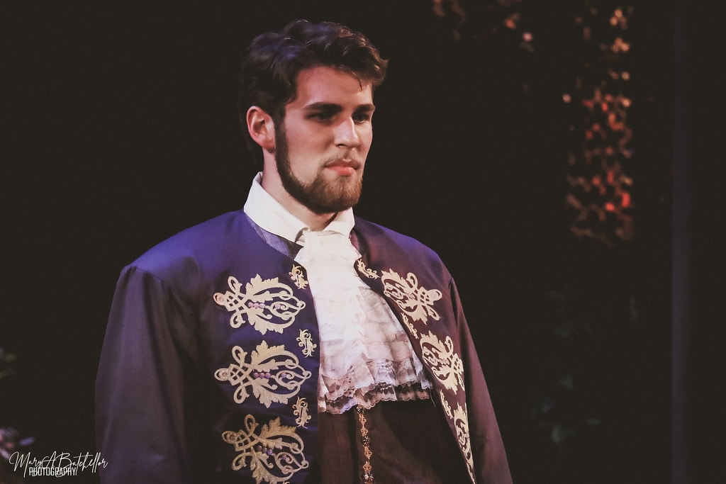 Into the Woods — Cinderella's Prince in a purple embroidered jacket stands in a dramatic close-up portrait