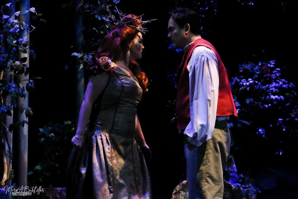 Into the Woods — the Act II Witch with red hair faces the Baker in his red vest in the purple-lit forest