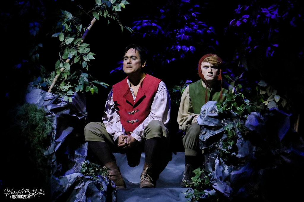Into the Woods — the Baker in a red vest and Jack in a green vest sit together on rocks in the blue-lit forest