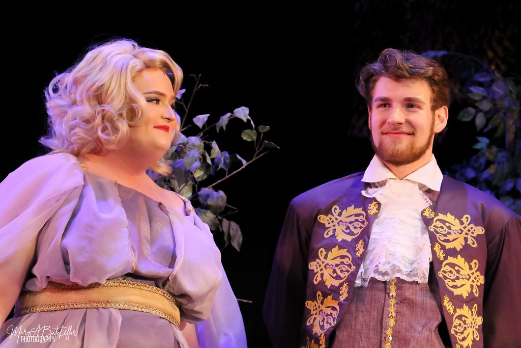 Into the Woods — Rapunzel in a white gown and Cinderella's Prince in a purple embroidered jacket share a close moment in the dark forest