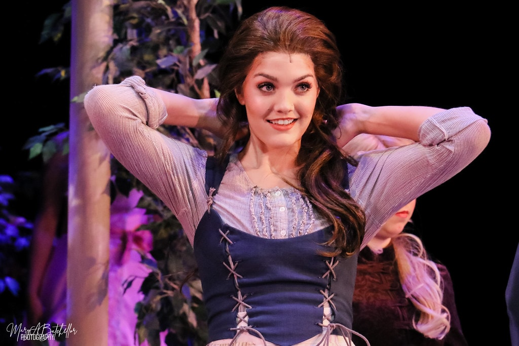 Into the Woods — the Baker's Wife in a blue lace-up corset dress smiles with hands behind her head in the forest