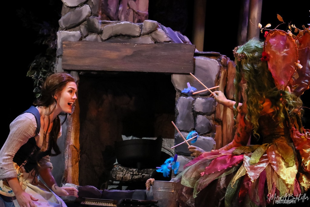 Into the Woods — the Baker's Wife reacts in shock as the Act I Witch holds blue bird puppets over a stone fireplace