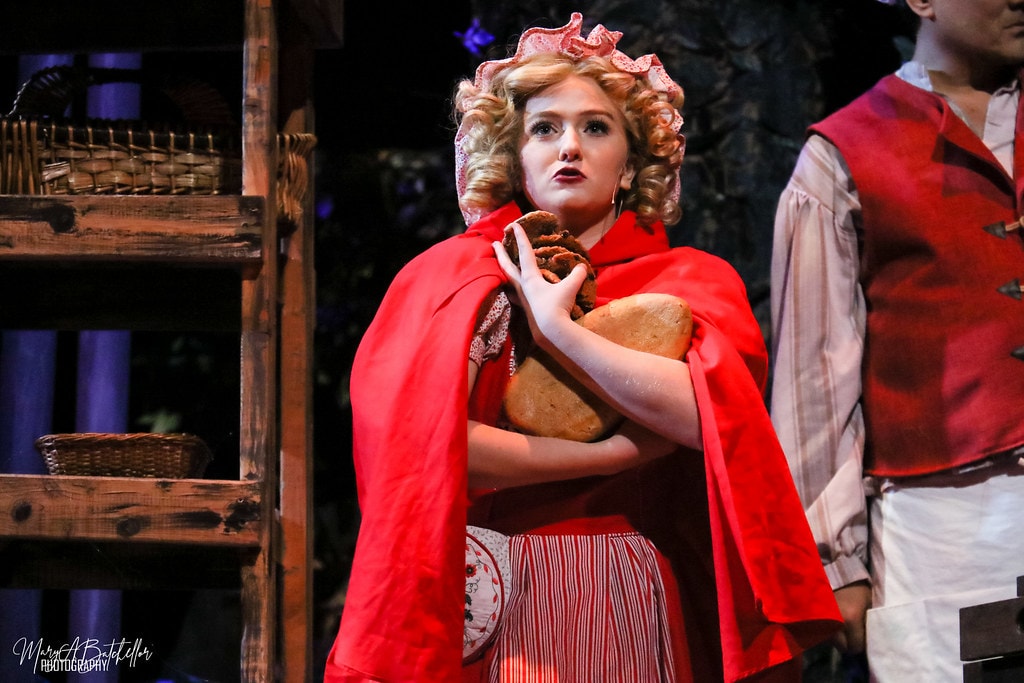 Into the Woods — Little Red Riding Hood in her red cape clutches a loaf of bread beside the Baker in the bakery set