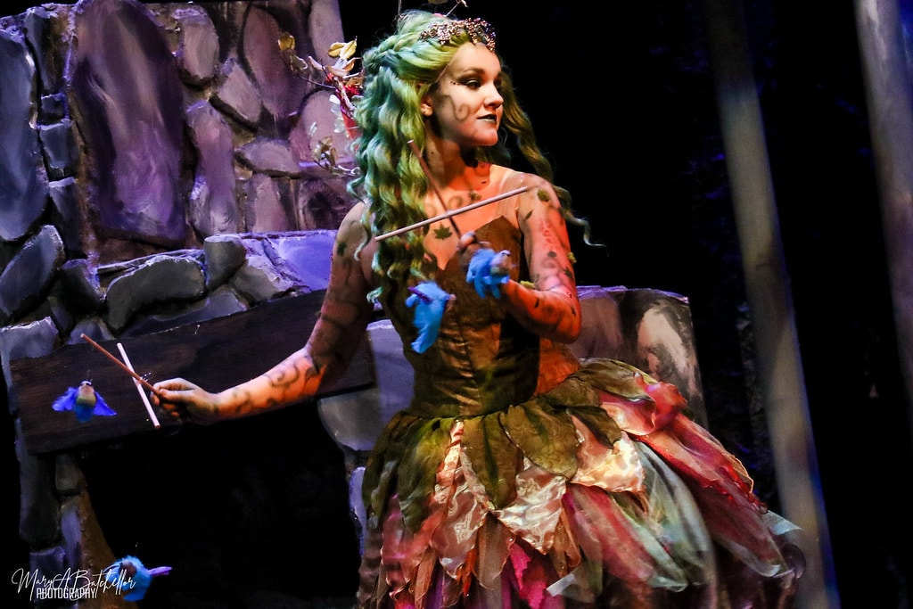 Into the Woods — the Act I Witch in a colorful leaf-and-petal gown holds blue bird puppets on wands in the stone fireplace scene