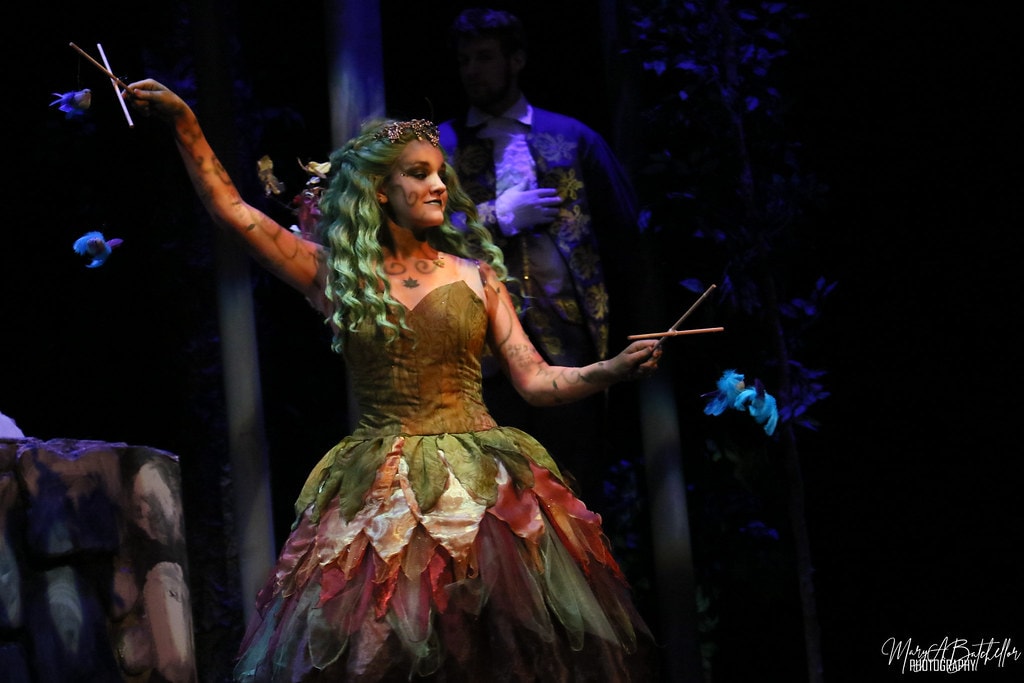 Into the Woods — the Act I Witch in a leaf-and-petal gown dances with arms outstretched as Cinderella's Prince stands behind her in the blue-lit forest