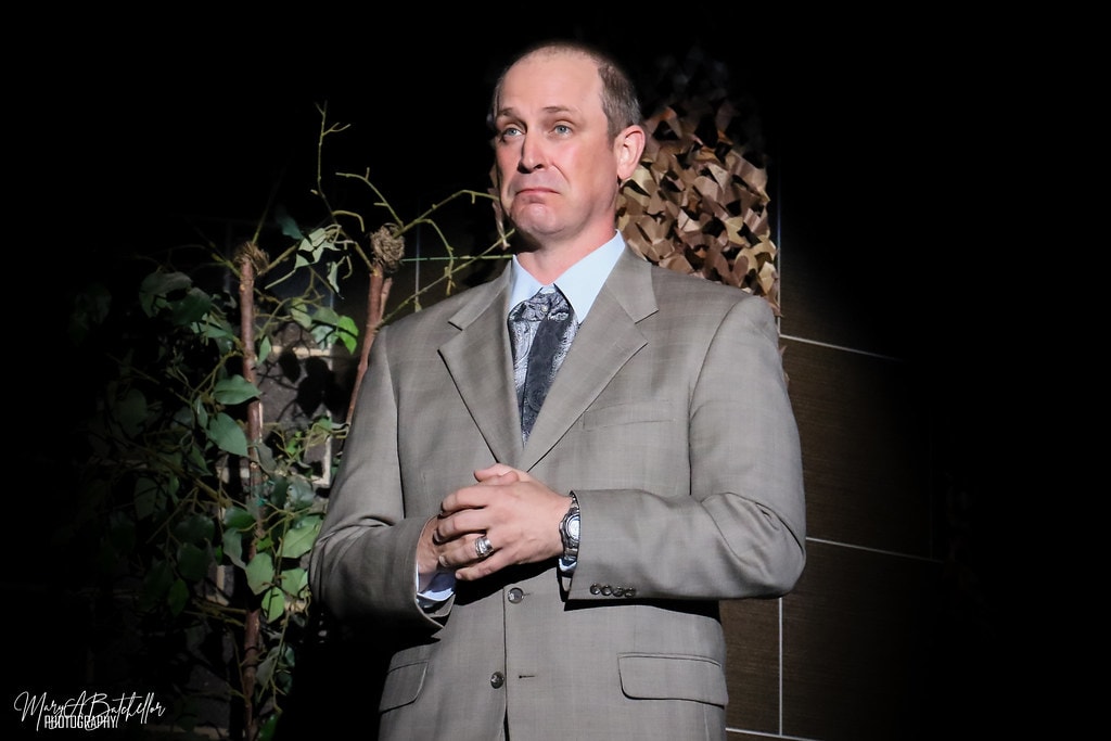 Into the Woods — the Mysterious Man in a grey suit stands alone in a dramatic spotlight before the dark forest backdrop