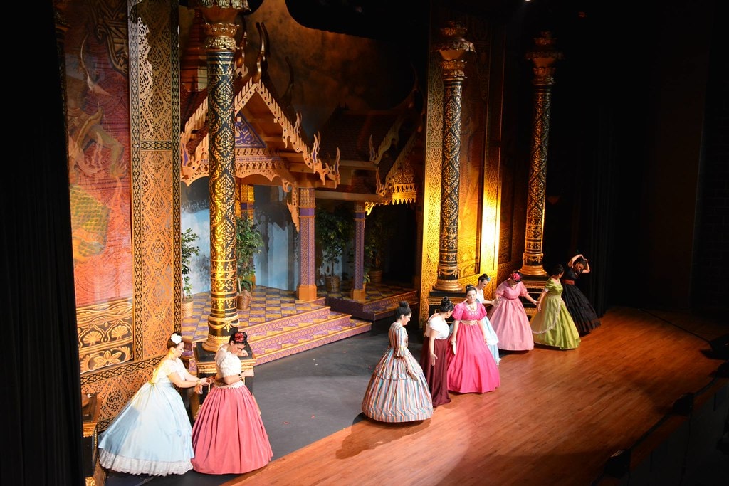 The King & I — women in Victorian ball gowns of pink, blue, and striped fabric dance in pairs across the warm golden stage floor before the ornate temple columns