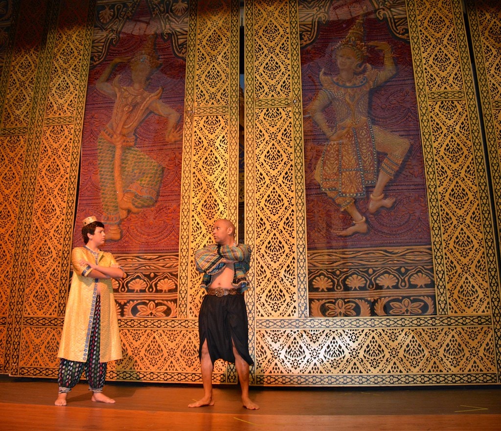 The King & I — two men in Siamese court costumes face each other before the towering golden Thai tapestry backdrop with classical dancer figures
