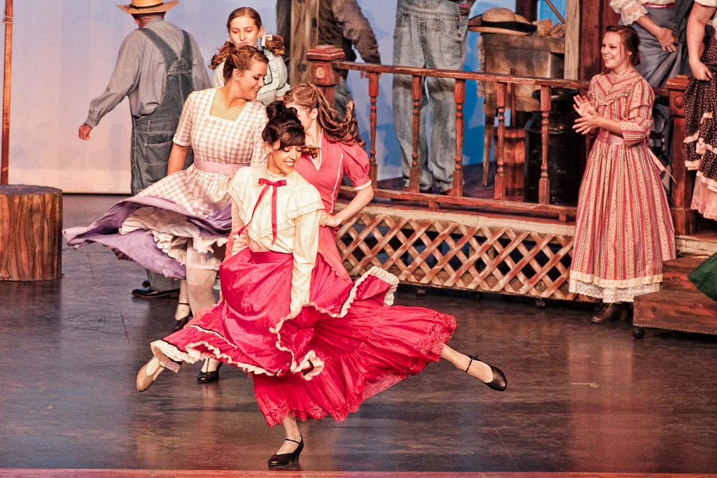 Oklahoma! — Laurey in a red ruffled skirt and white blouse executes a sweeping dance kick at center stage as two women in prairie dresses spin behind her and a woman in a striped dress watches from the porch railing before the farmhouse set