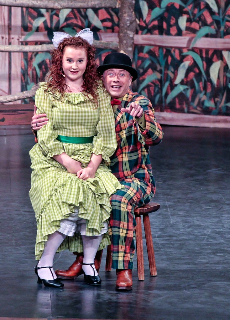 Oklahoma! — Ado Annie in her green gingham dress and white bow sits on a stool beside Ali Hakim in his colorful plaid suit and black bowler hat, who points outward with a broad grin before the painted cornfield backdrop