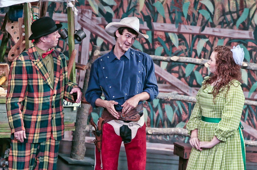 Oklahoma! — Ali Hakim in his plaid suit at left, Curly in his blue snap-button shirt and red chaps at center, and Ado Annie in her green gingham dress at right share a three-way scene before the painted cornfield fence backdrop