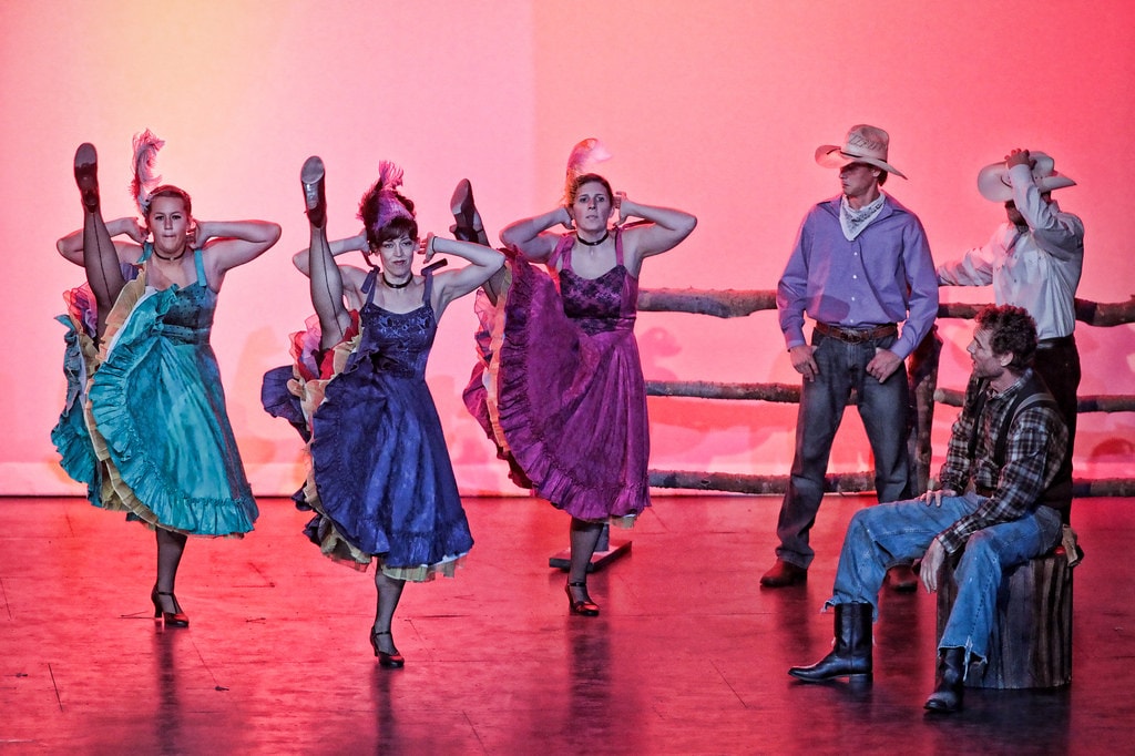 Oklahoma! — the dream ballet sequence: three women in jewel-toned saloon dresses — teal, navy, and purple — kick their heels high in a can-can line before a pink-lit backdrop as two cowboys watch from the fence at right