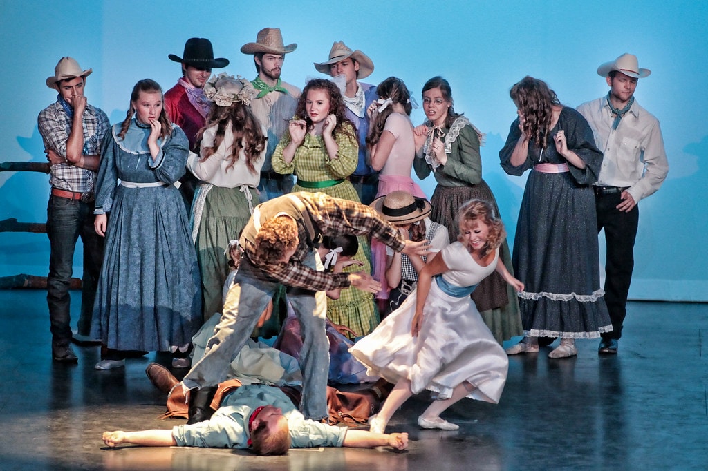 Oklahoma! — the dramatic finale confrontation: over 15 cast members in period costumes crowd the blue-lit stage as Laurey in her white dress reaches toward the fallen Jud Fry on the floor while Will Parker bends over him and the ensemble looks on in shock