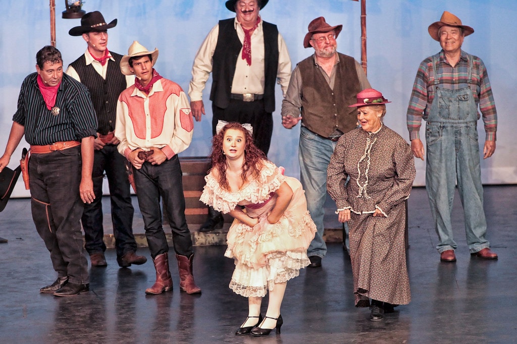 Oklahoma! — Ado Annie in her cream lace ruffled dress curtsies at center stage as Aunt Eller in her brown calico dress and pink hat stands beside her, flanked by four cowboys in western shirts and hats and two farmers in overalls before the pale blue sky backdrop
