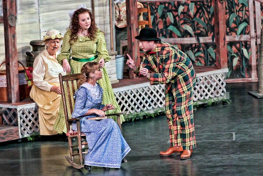 Oklahoma! — Aunt Eller in her cream blouse and bonnet sits in a rocking chair as Laurey in blue calico stands behind her and Ali Hakim in his loud plaid suit leans forward pointing at Laurey on the farmhouse porch set