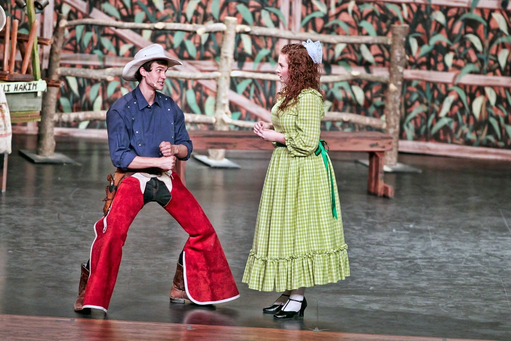 Oklahoma! — Curly in his white cowboy hat and red chaps faces Ado Annie in her green gingham dress in a tense confrontation before the cornfield fence backdrop