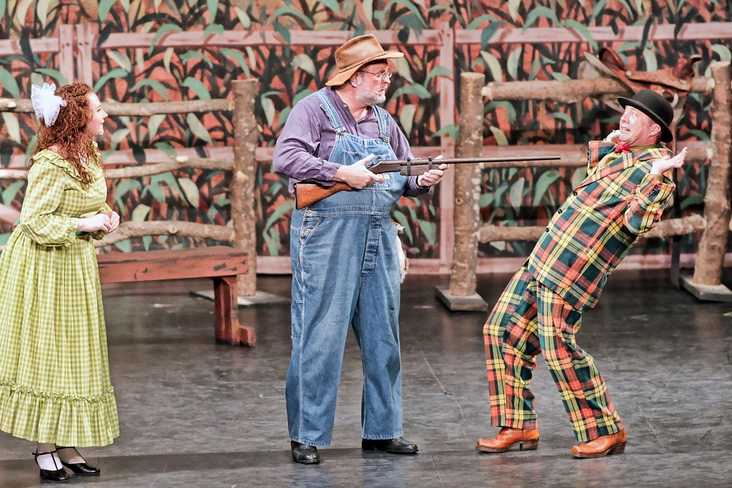 Oklahoma! — Ado Annie in her green gingham dress watches as Aunt Eller's husband in overalls levels a shotgun at Ali Hakim in his plaid suit, who recoils in comic fright before the cornfield fence backdrop