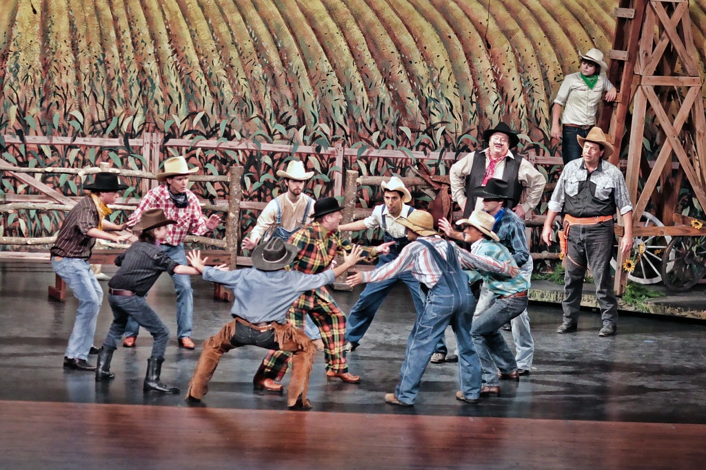Oklahoma! — a large ensemble of cowboys in hats and work clothes squares off in a brawl on the barn dance set, with Jud Fry in his black hat at center and Curly in his chaps visible among the crowd before the golden wheat field backdrop