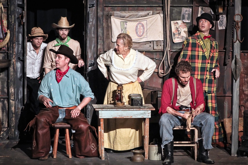 Oklahoma! — six cast members in the smoky barn interior set: Curly in his teal shirt and leather chaps sits at a stool at left as Aunt Eller stands center with hands on hips, Jud Fry in his red shirt sits at right, and three cowboys peer through the barn door behind them