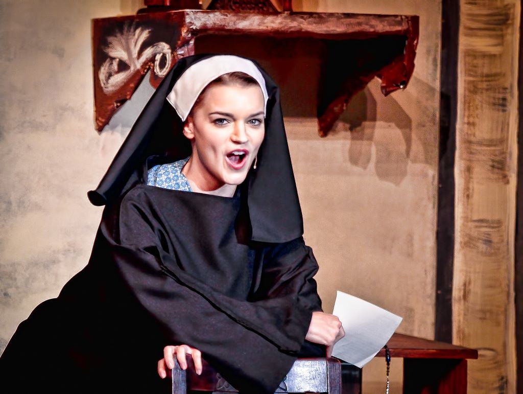 The Sound of Music — a young nun in a black habit leans forward with wide eyes and an open mouth, holding a letter, in a dramatic comedic scene before the convent set