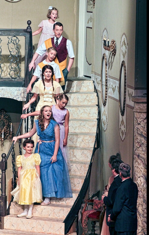The Sound of Music — the seven von Trapp children in colorful formal dresses and suits descend the grand ornate staircase of the von Trapp villa set in a line