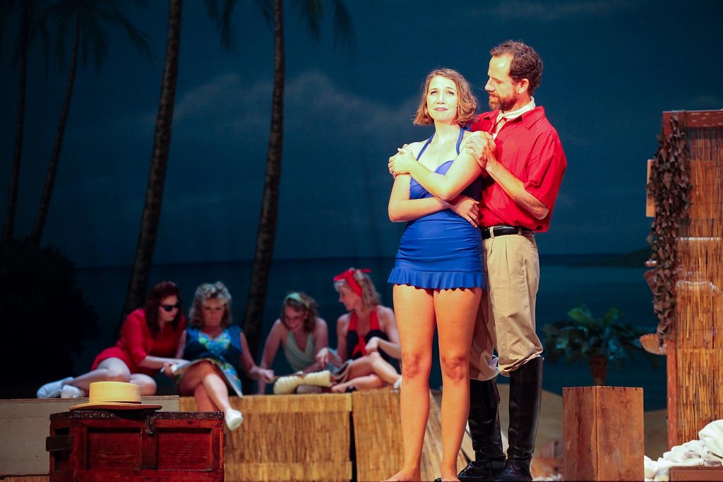 South Pacific — a young woman in a blue swimsuit stands in the arms of a man in a red shirt before a tropical beach backdrop, nurses and ensemble seated in the background