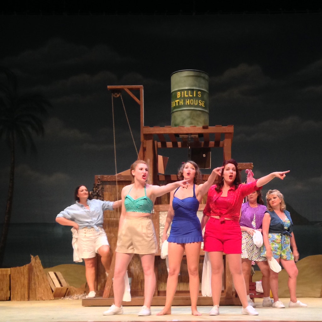 South Pacific — six women in colorful 1940s beach outfits sing and point dramatically before the Billis Bath House set against a dark sky backdrop