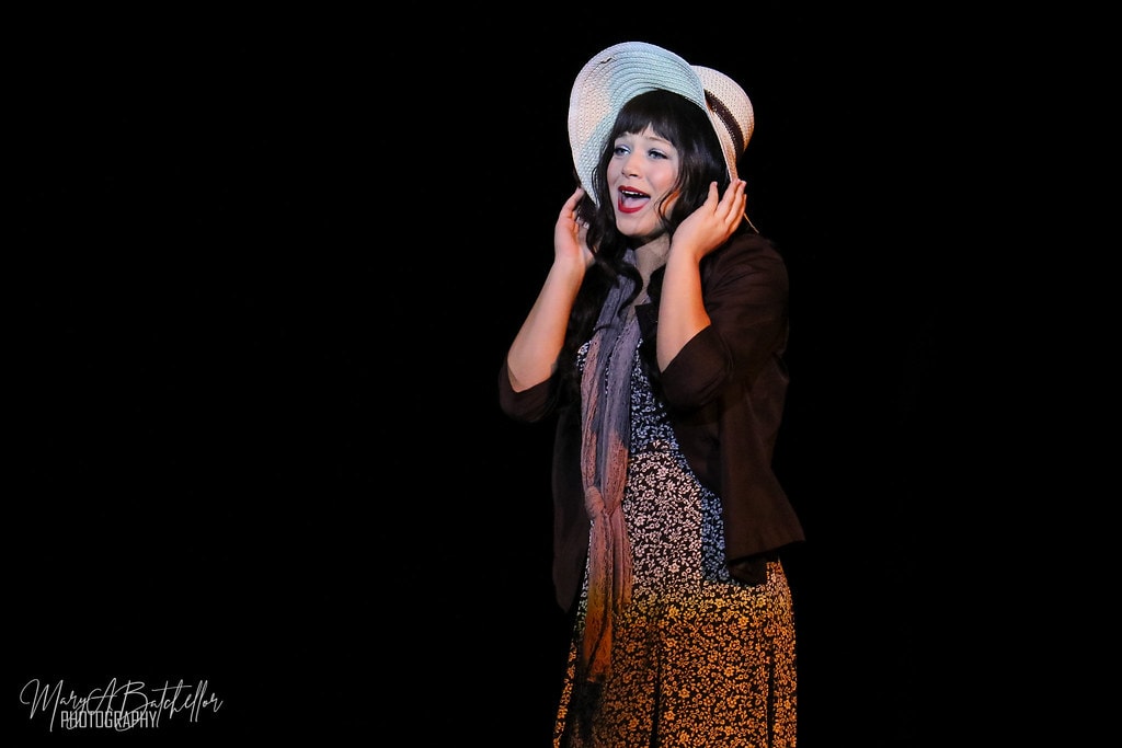 Thoroughly Modern Millie — Millie in a floral dress and brown cardigan holds her straw hat and sings a solo